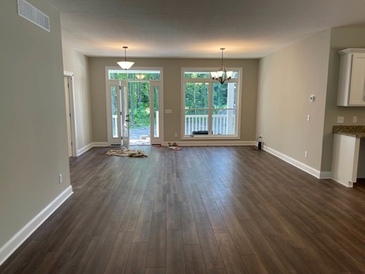 Open living room with rich hardwood flooring, white walls, large window overlooking porch, and modern light fixture.