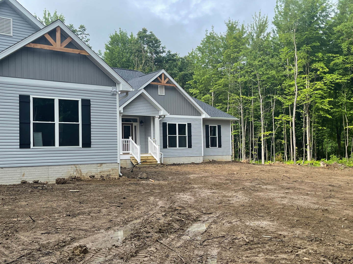 Two-story cottage-style home with white siding, black shutters, white framed windows, and a white staircase and railing leading to a muddy yard, surrounded by trees and dirt.