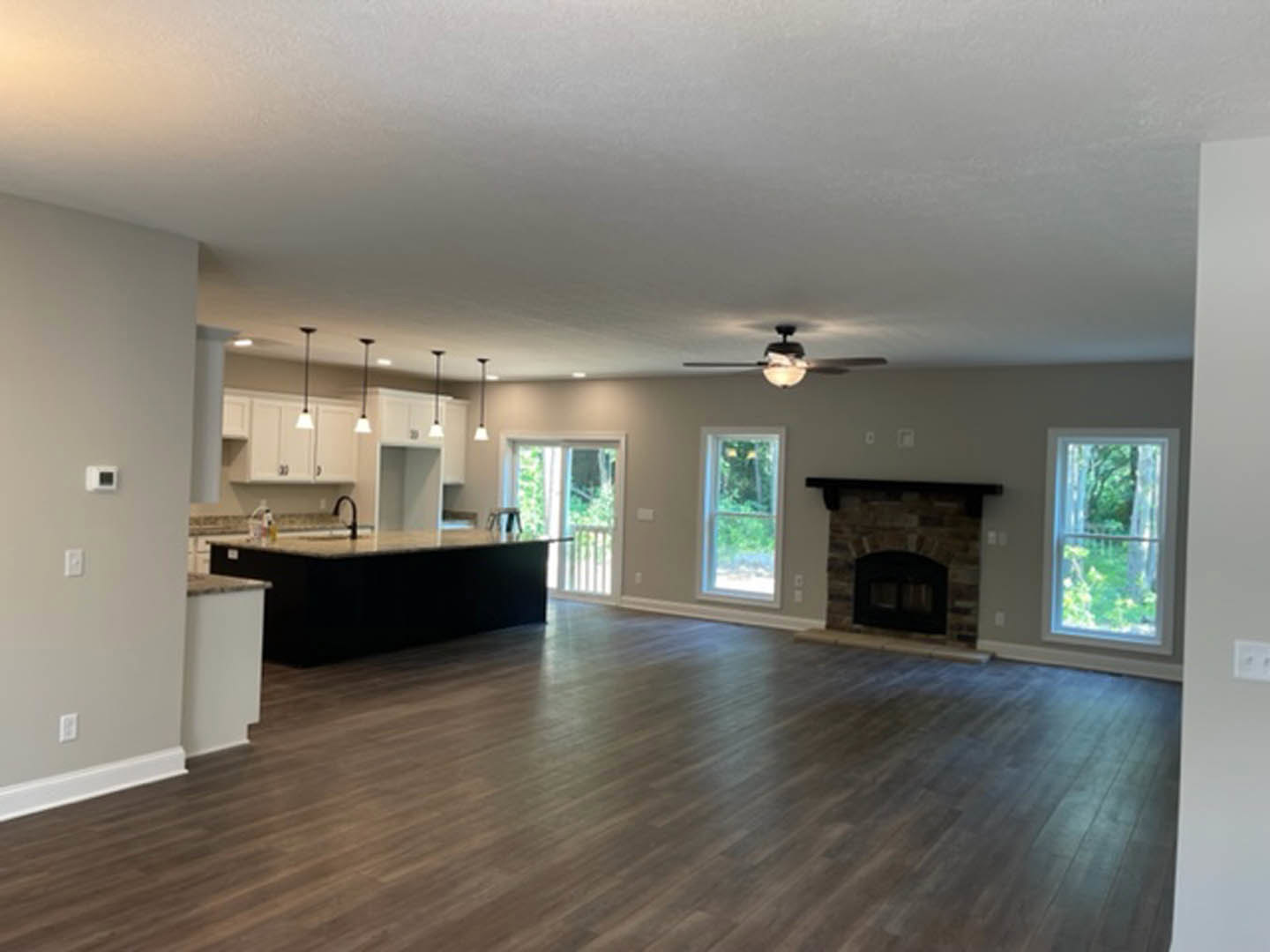 Spacious open floor plan featuring hardwood floors, modern kitchen with white cabinetry, central fireplace with black door, large windows offering outdoor views, and ceiling fan
