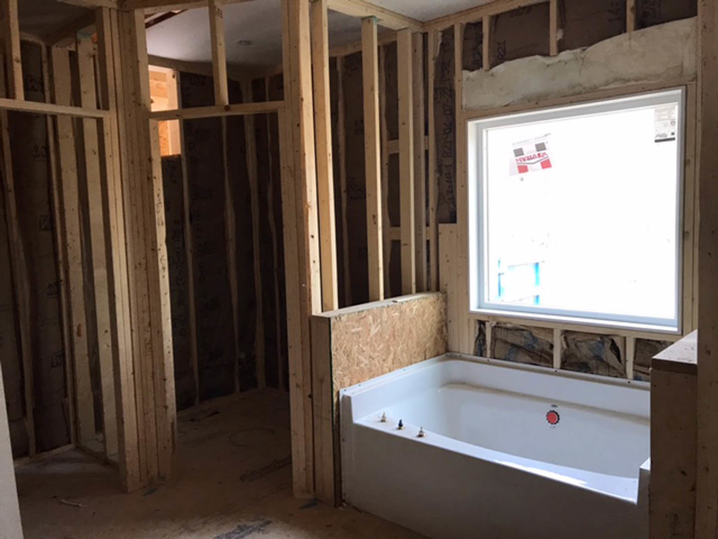 Bathroom under construction with exposed wooden framing, white bathtub featuring a red marking, unfinished plaster walls, and window with sticker.