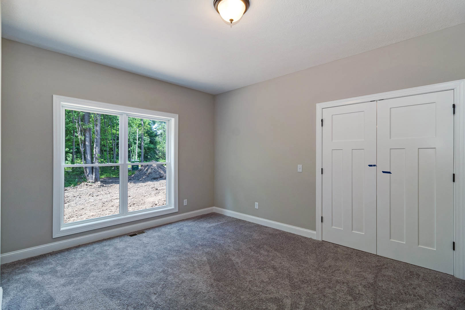 Bedroom with white closet doors featuring blue handles, large window overlooking trees, beige carpet flooring, and modern ceiling light fixture