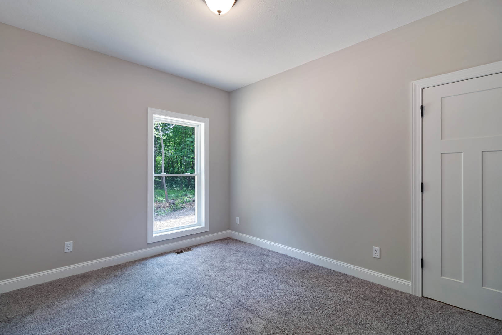 Bedroom with beige carpet, white paneled door, large window overlooking trees, white walls, ceiling light fixture, and crown molding
