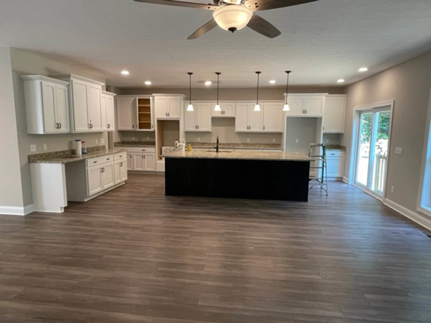 Spacious kitchen featuring a large central island with black countertop, wooden flooring, white cabinetry, stainless steel appliances, and ceiling fan; windows provide natural
