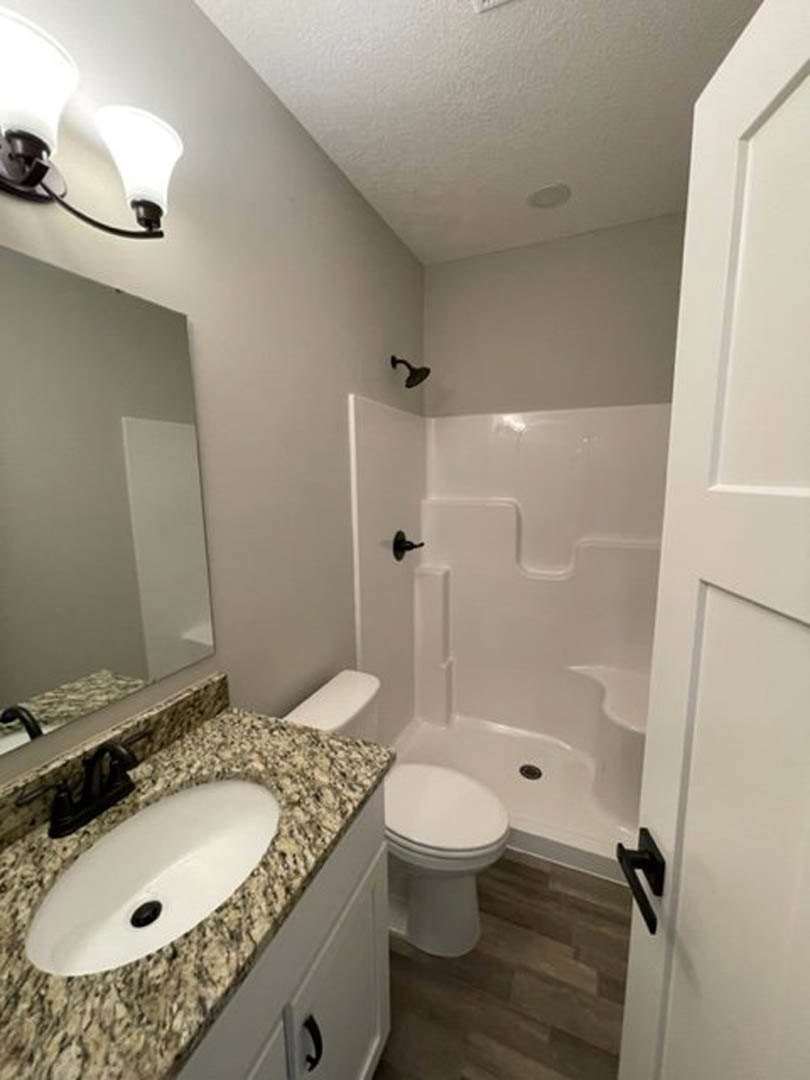 White oval sink with black drain set in a marble countertop, glass-enclosed shower with tile walls, modern toilet, brushed nickel fixtures, and light gray tile flooring