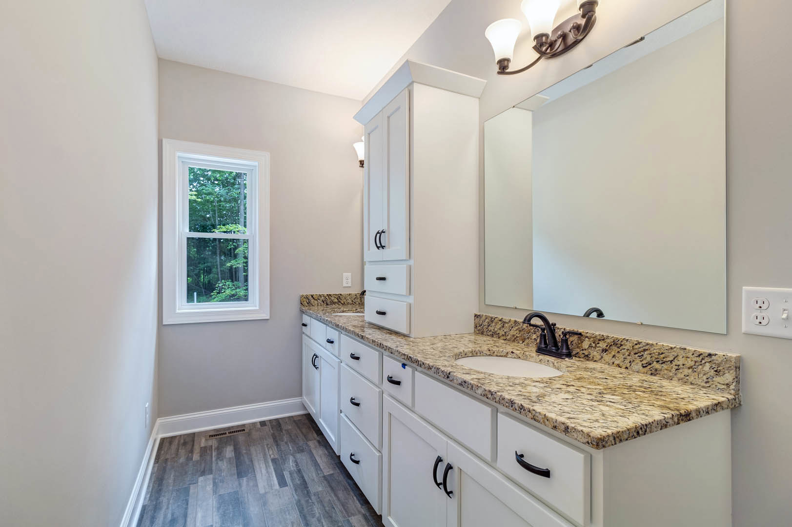 Bathroom with marble countertop, rectangular mirror above sink, chrome faucet, wood flooring with floor vent, window showing trees outside, and modern light fixture.