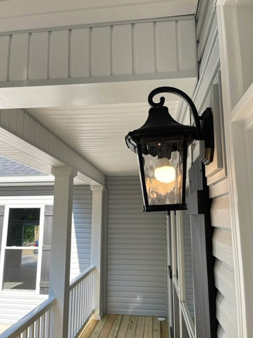 Black metal lantern-style wall sconce mounted beside white porch railing, wood handrail, and balusters, with exterior siding and outdoor space visible.