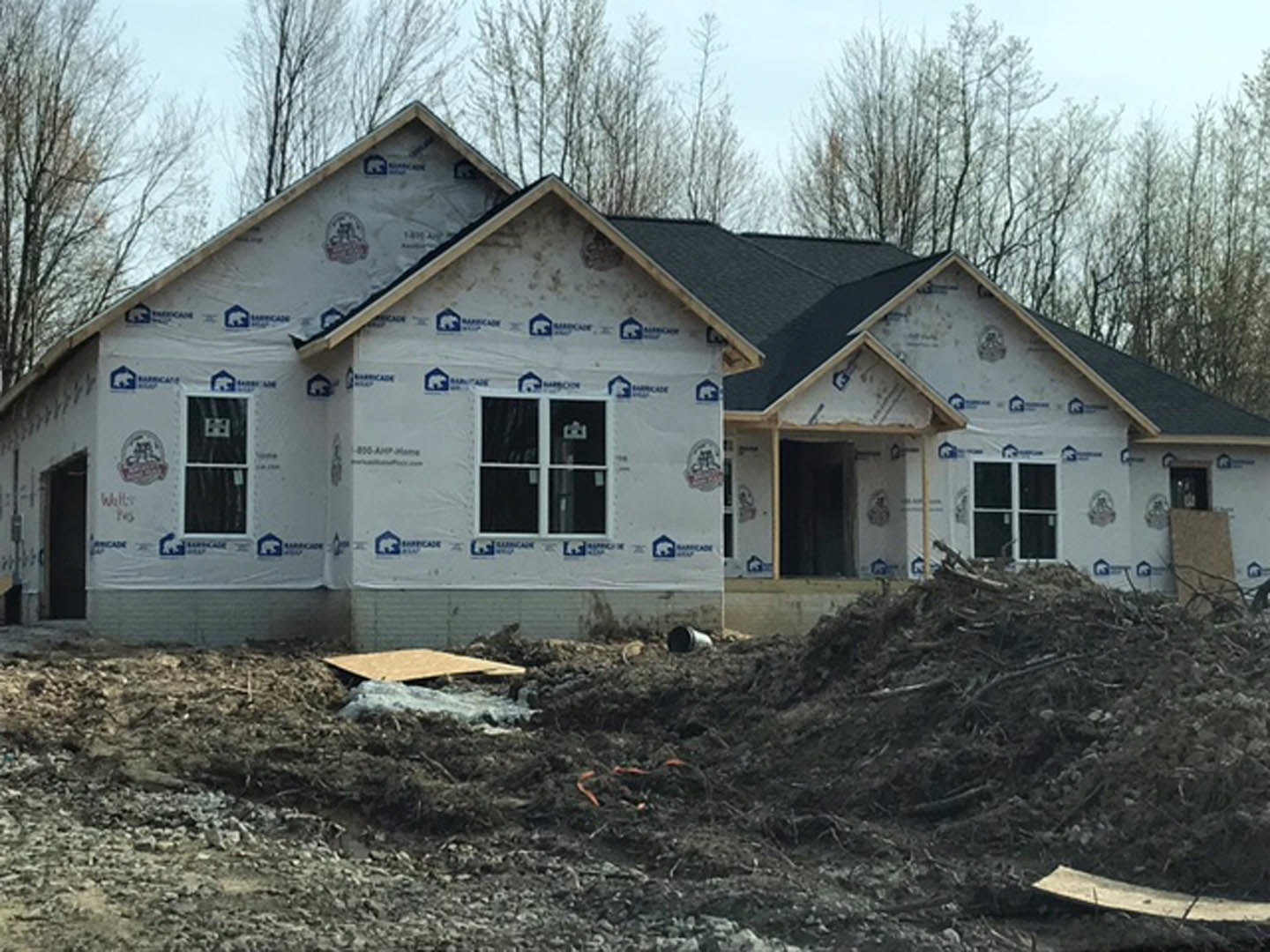 Two-story house under construction with exposed framing, white plastic sheeting, piles of dirt and debris in foreground, surrounded by trees under a clear sky