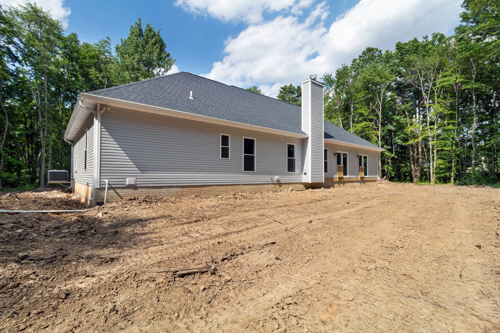 White custom home with large window and white trim, surrounded by dirt lot and mature trees under cloudy sky