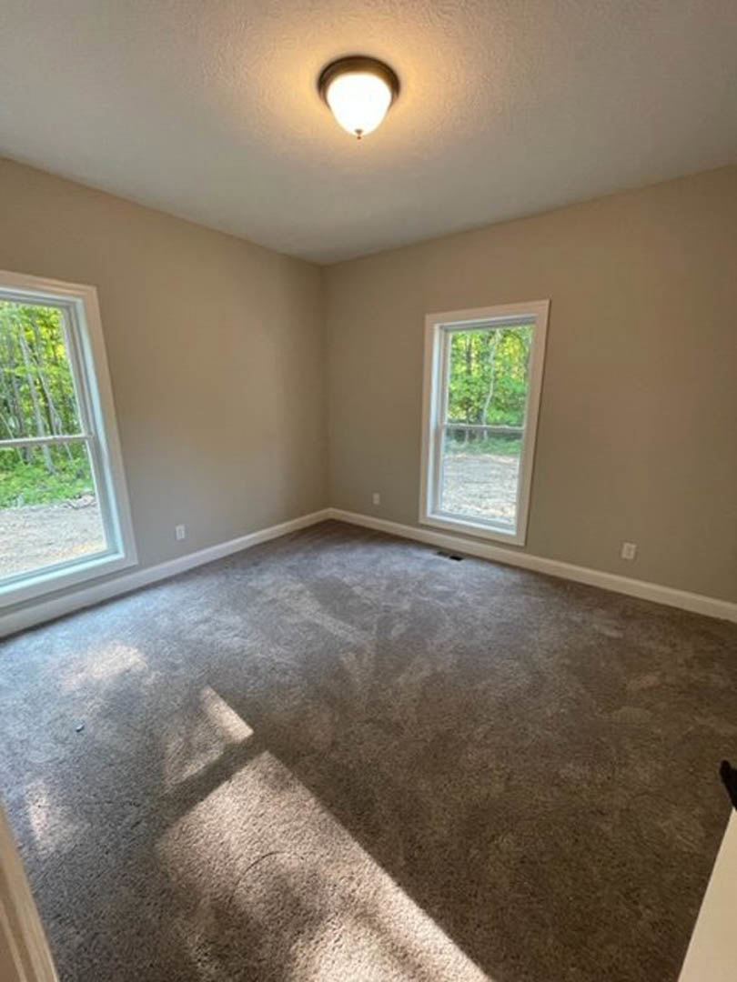 Carpeted room with large windows overlooking trees, white plaster walls, ceiling-mounted light fixture, and laminate flooring near the entry.