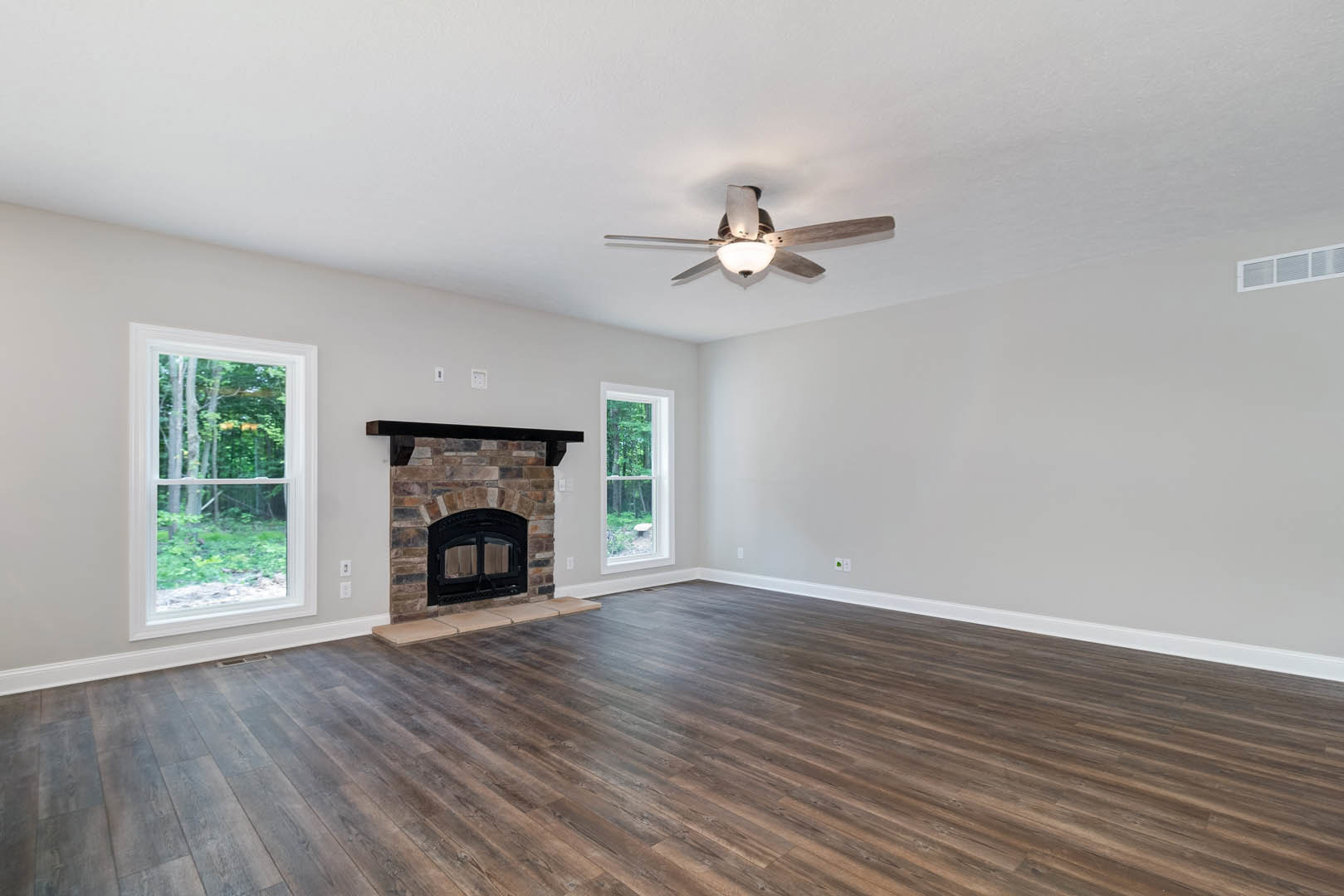 Living room with wood flooring, brick fireplace featuring a black metal-framed glass door, black ceiling fan with light, and large window overlooking trees.