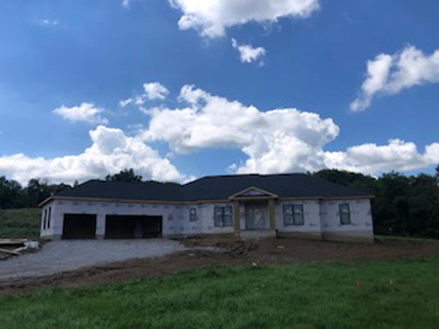 Modern cottage-style house under construction with black roof, large windows with black frames, surrounded by green grass and mature trees under a cloudy sky