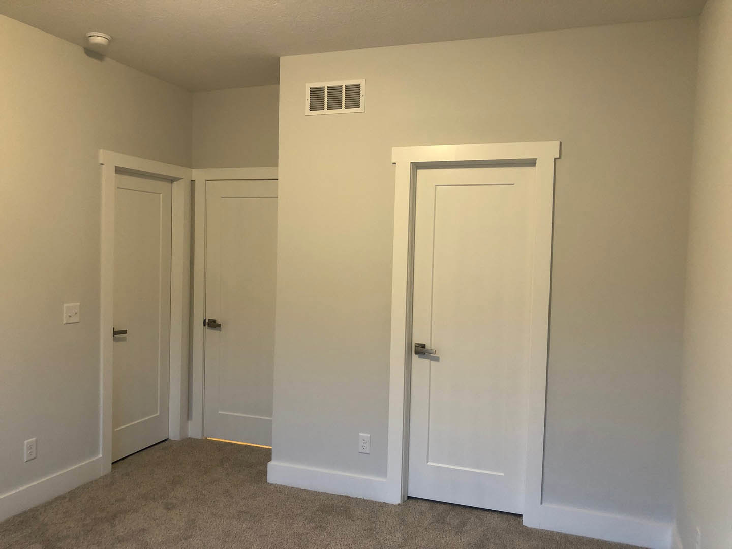 White paneled door with silver handle, beige carpet flooring, white plaster walls, and a rectangular air vent near the baseboard