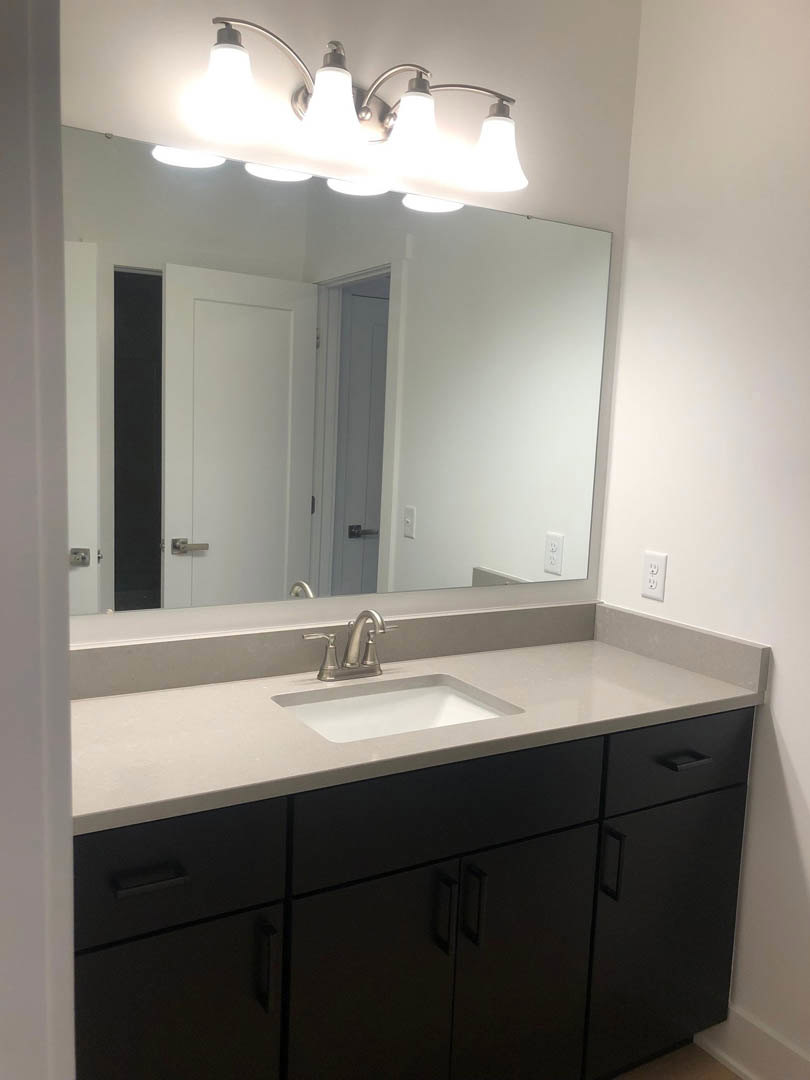 White quartz countertop with undermount sink, chrome faucet, rectangular wall mirror, four-bulb vanity light, light gray tile backsplash, white cabinetry