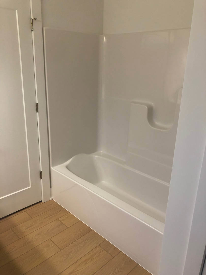 Freestanding white bathtub beside light wood flooring, white walls, and white door with metal hinge in modern bathroom