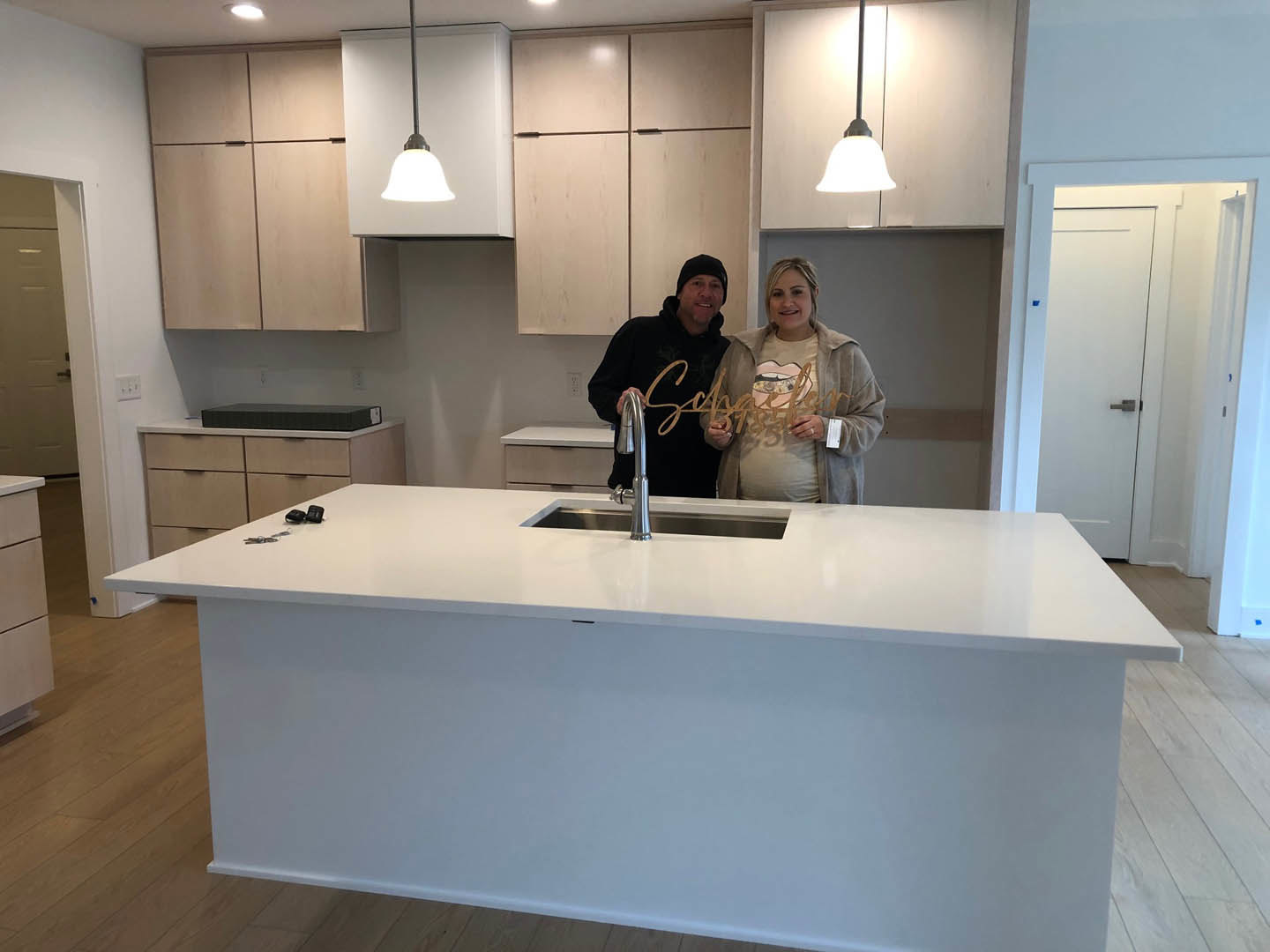 Couple standing in modern kitchen with white countertops, light cabinetry, stainless steel sink, and wood flooring