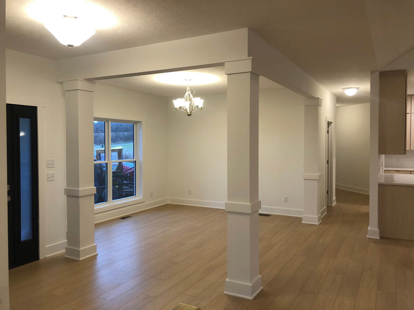 Open room with white pillars, wood laminate flooring, ceiling light fixture with five bulbs, blue rectangular object near wall, window showing tractor parked outside