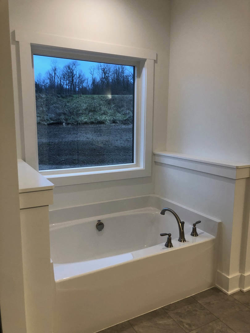 Freestanding bathtub beneath large window overlooking field of trees, chrome faucet, light tile flooring, white walls