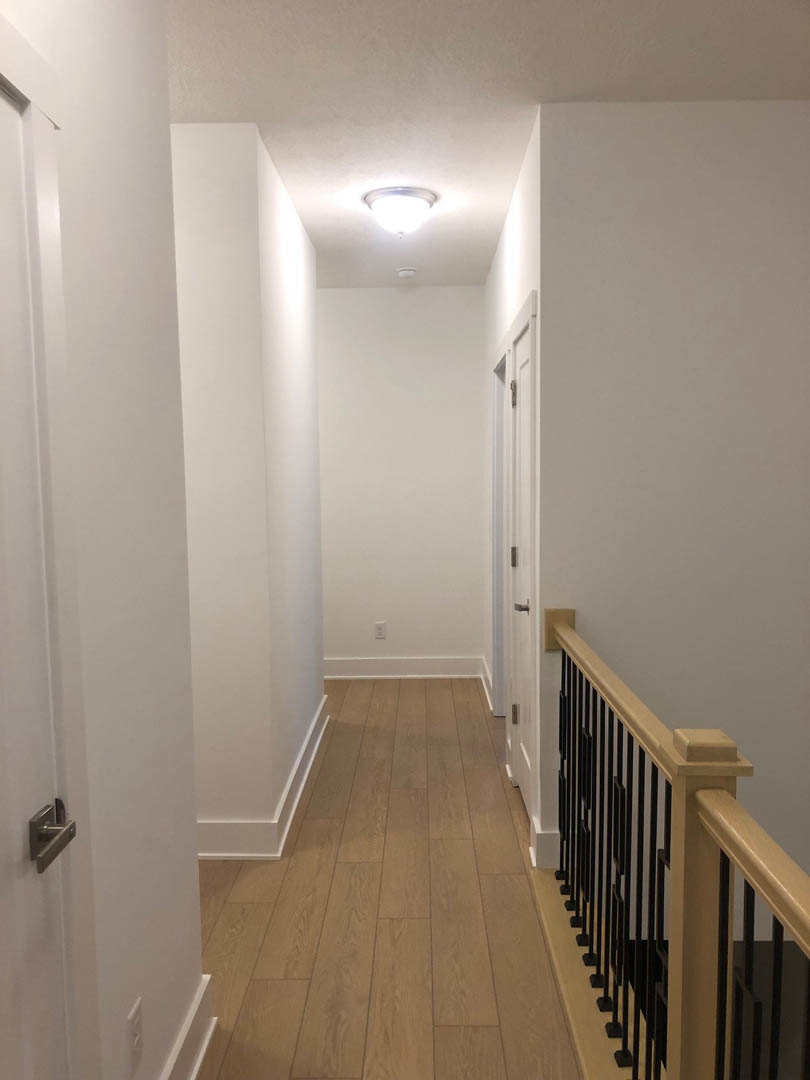 Hallway with smooth white walls, warm wood flooring, white pillar, modern handrail, close-up of a brushed metal door handle, and ceiling light fixture with circular trim