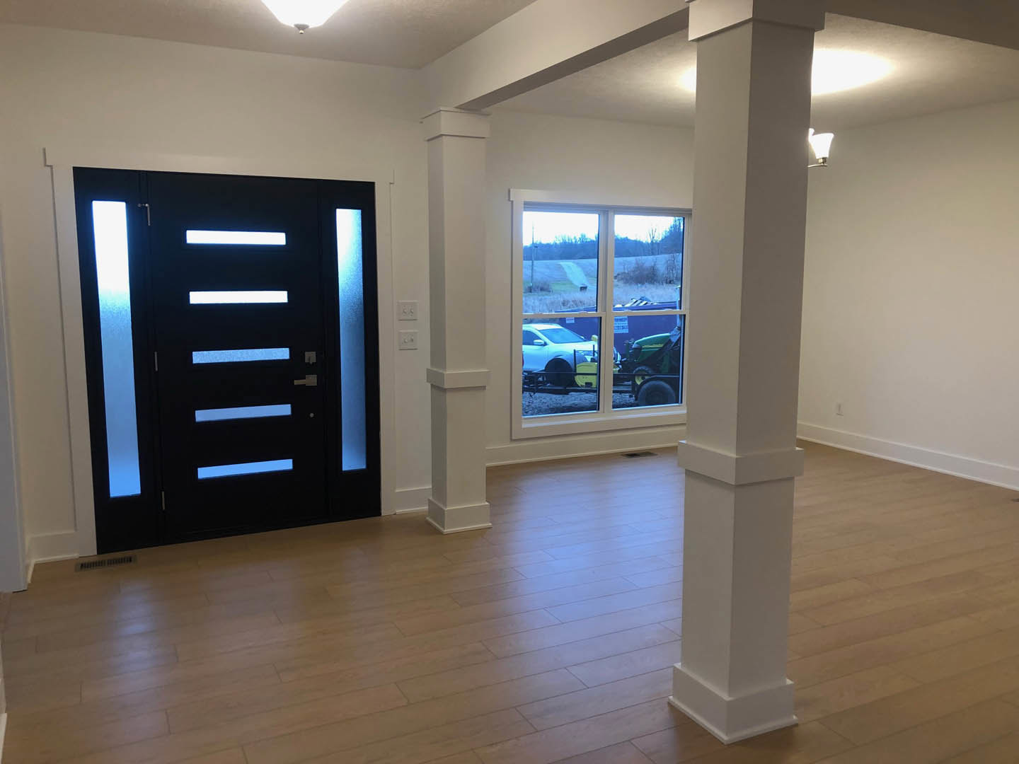 Black door with blue accent lights, wood laminate flooring, white pillar, window overlooking a field with a tractor, neutral walls.
