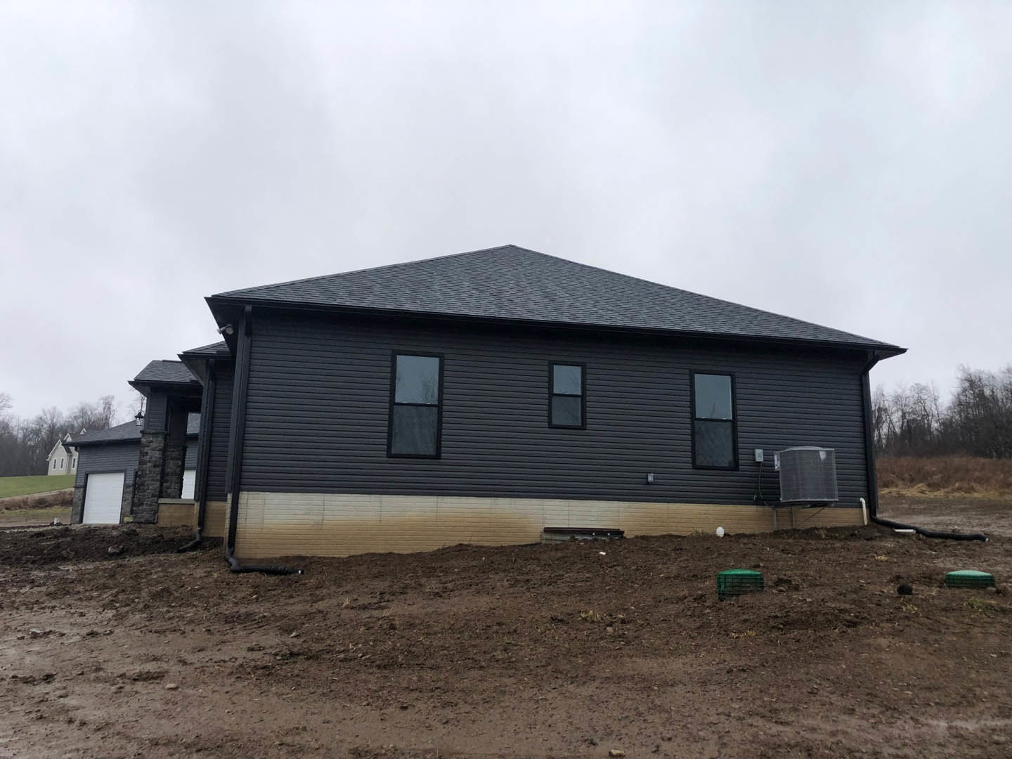 Black siding house with square-paned black frame windows, dirt hill in foreground, green container on ground, close-up of outdoor heater, cloudy sky overhead.