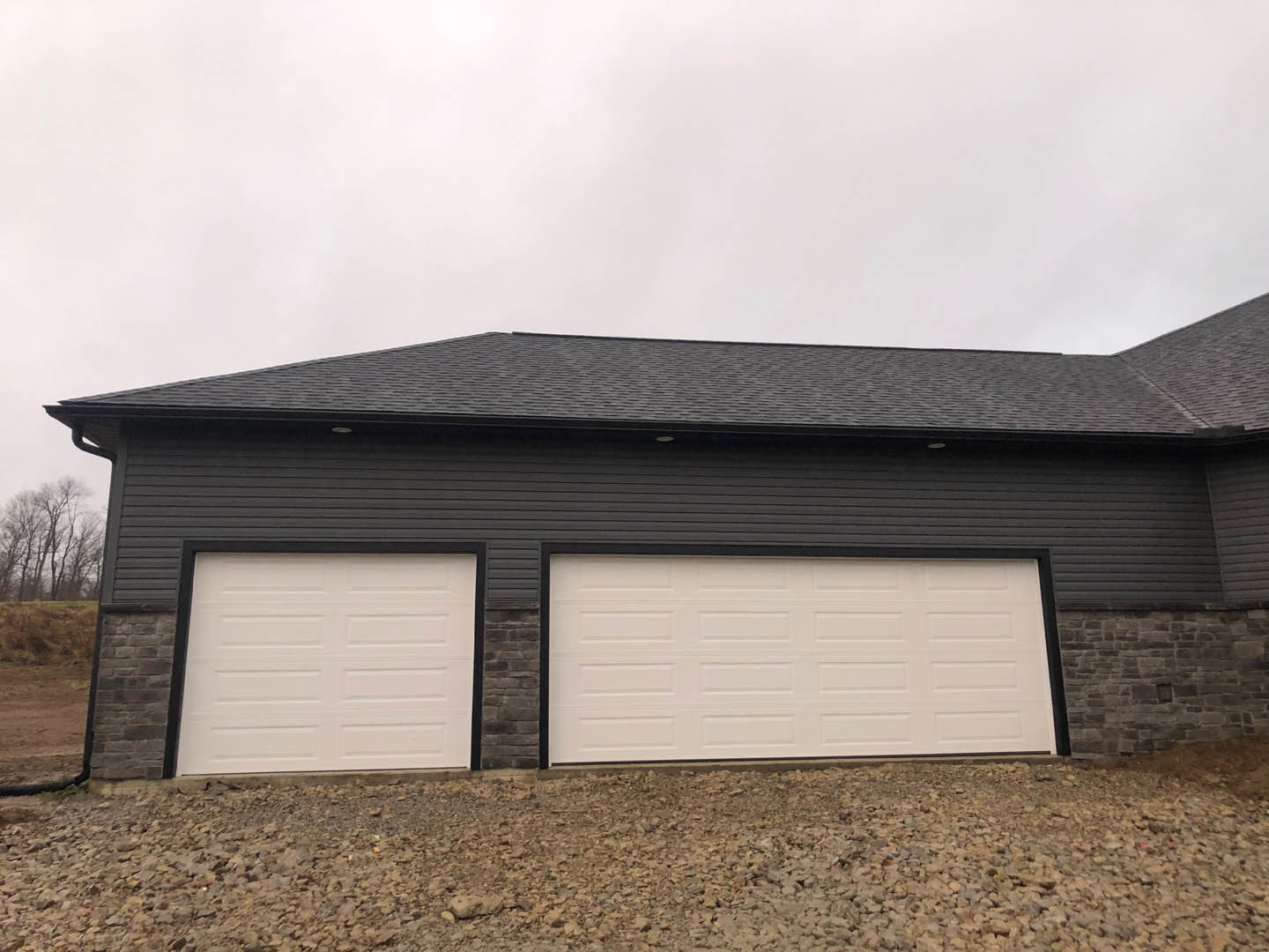 White garage with two black-trimmed doors, stone pile in front, leafless trees and grassy field in background, gray roof visible above.