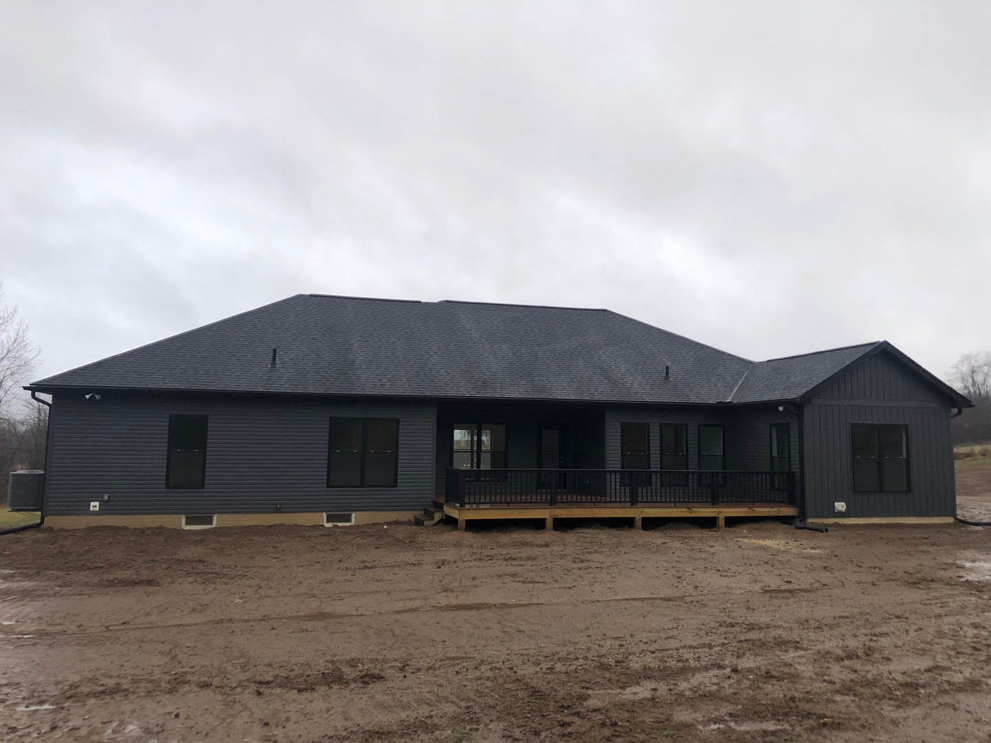Two-story home with wide covered porch, white siding, black-framed windows, wood deck with railing, gabled roof, dirt yard with tire tracks, scattered trees in background