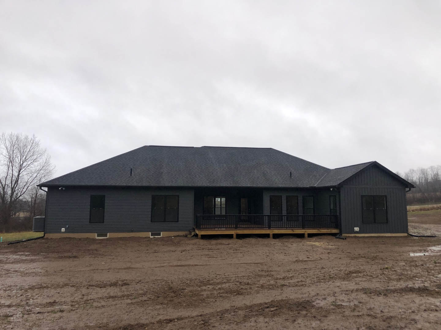 White custom home with expansive covered porch, black-framed windows, metal railing, surrounded by dirt lot and mature tree in background