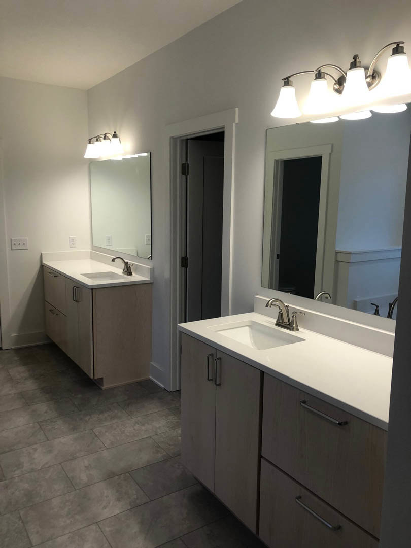 Double vanity bathroom with white cabinets, two undermount sinks, wide framed mirror, four-light fixture above, white walls, and partial view of a door.