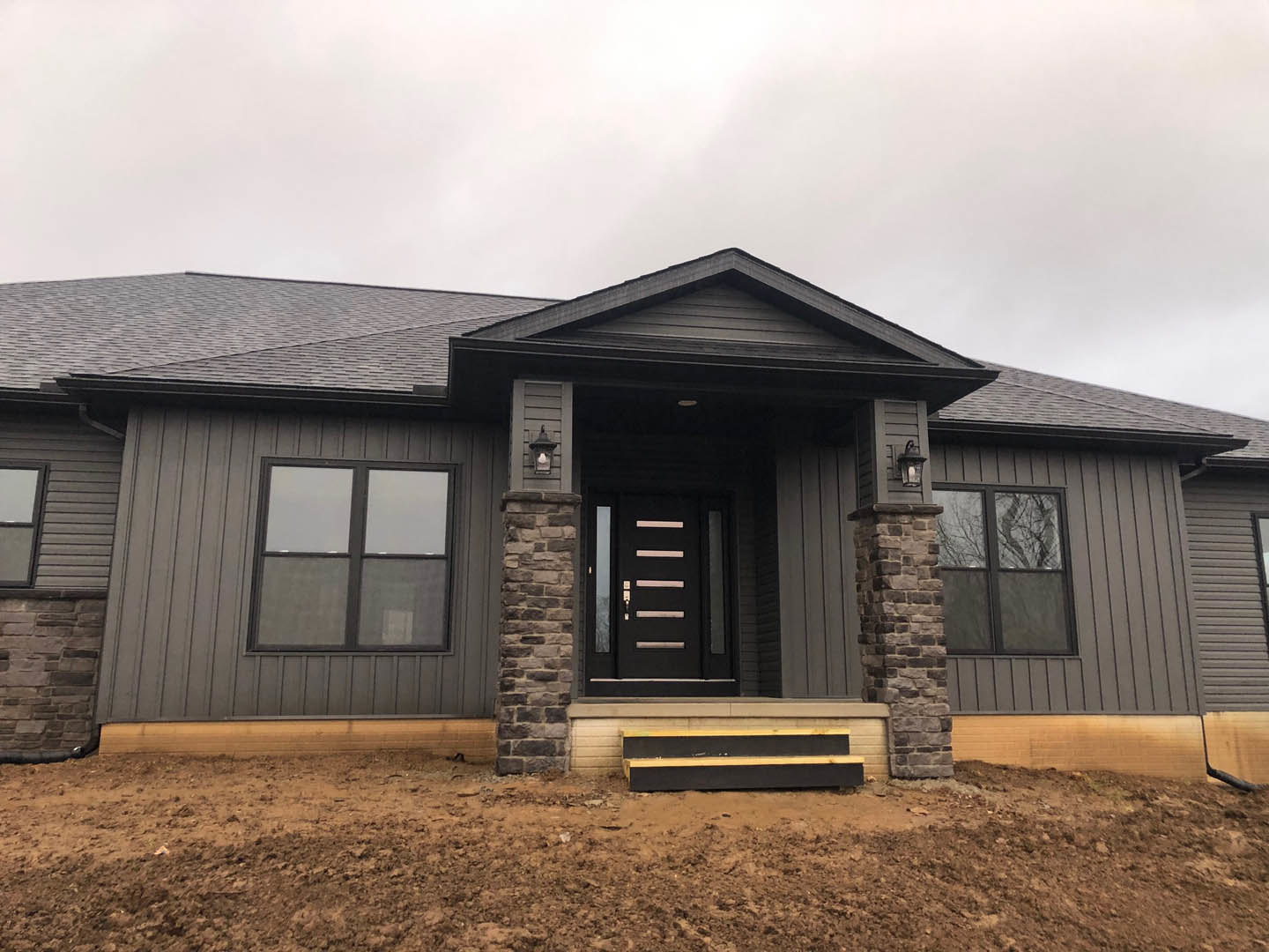 White siding house with black front door, covered front porch, large windows, and dirt landscaping under a cloudy sky