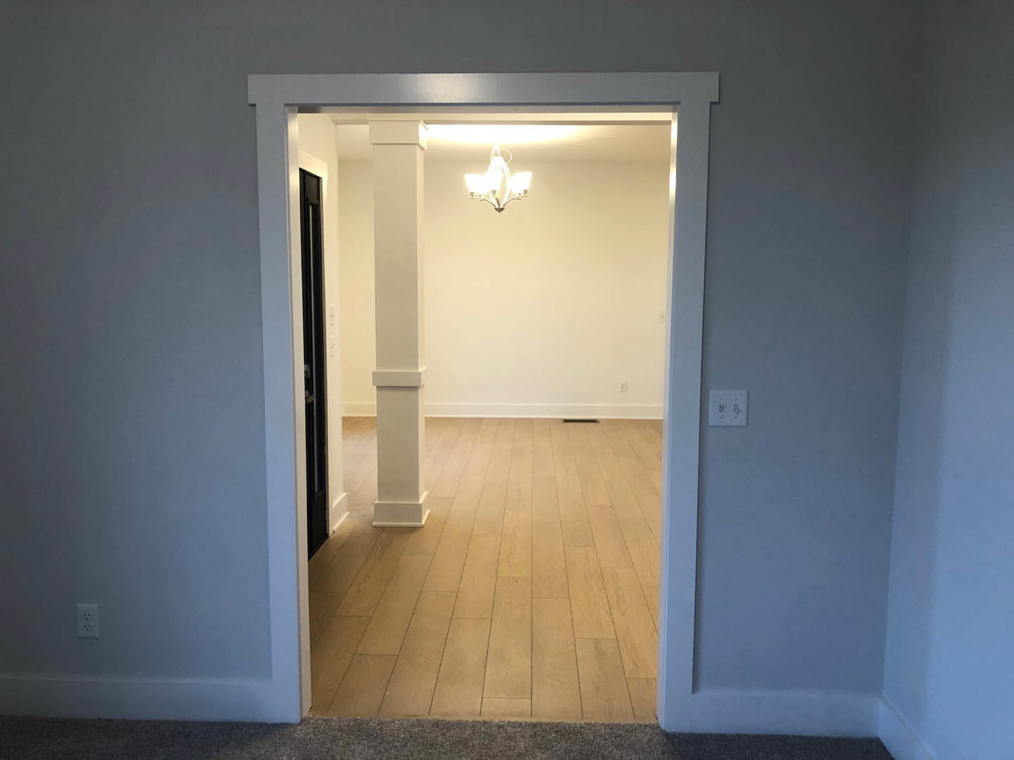 Wide doorway framed by white pillars opens to a room with wood flooring, white walls, and a modern chandelier; white light switch with black border visible on wall.