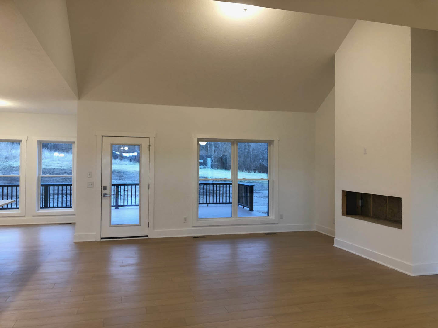 Living room with wood flooring, plaster walls, central fireplace, glass doors opening to snowy landscape, large windows with ocean view, metal railing visible outside