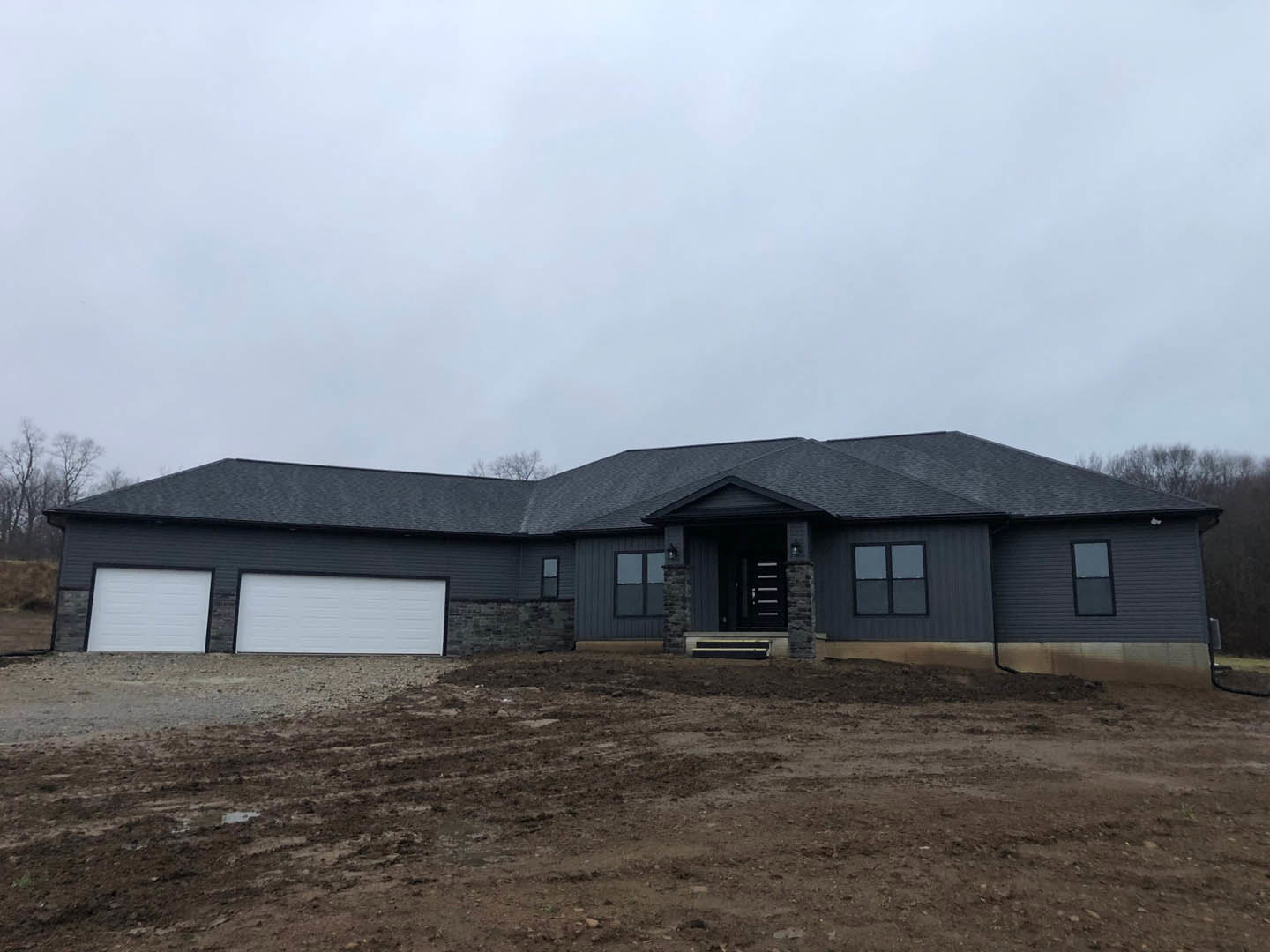 Modern house with white walls and black trim, stone accent wall, covered porch, large square windows, surrounded by dirt field and sparse trees under cloudy sky