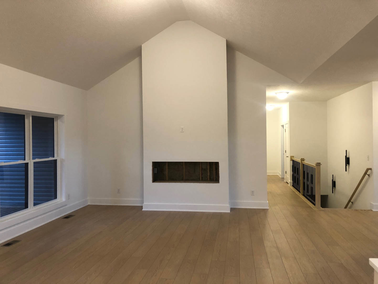 Living room with wide plank hardwood flooring, white plaster walls, built-in fireplace, and large window; white electrical outlet visible on wall, metal railing in foreground