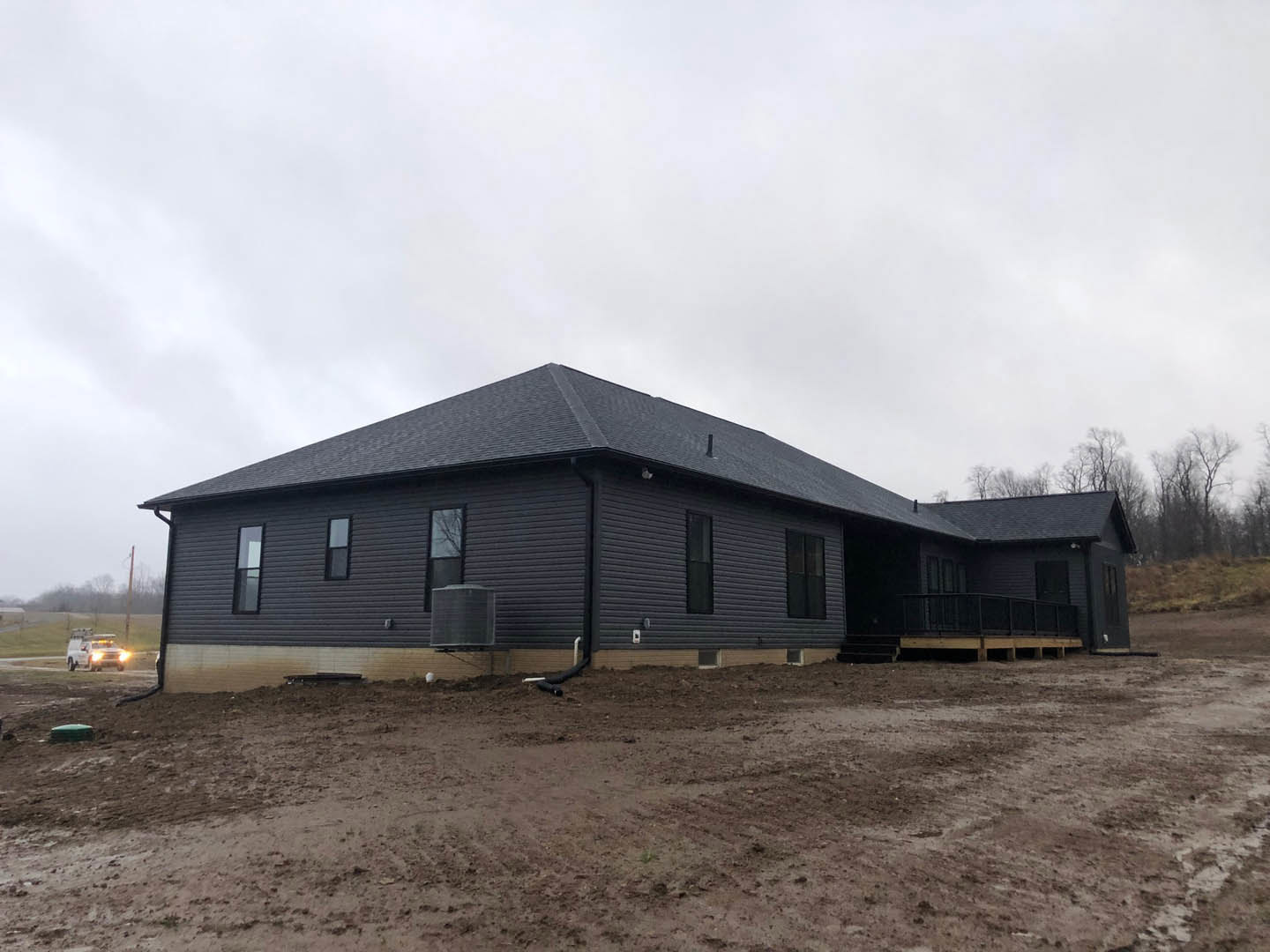 Two-story house with black roof, expansive covered porch, and spacious wooden deck; white pickup truck parked on dirt lot, black metal container nearby, cloudy sky overhead