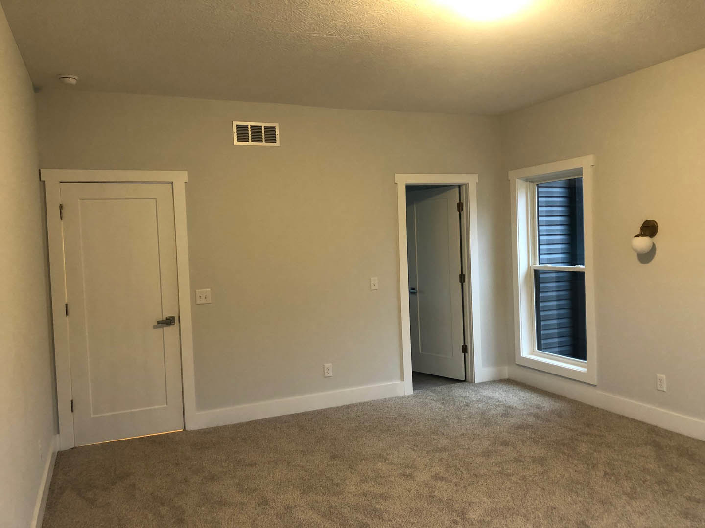 Bedroom with light gray carpet, white walls, two white doors with black handles, white-framed window, wall vent, and smooth plaster ceiling