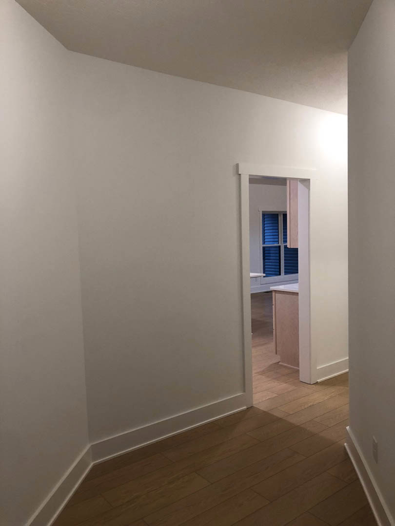 Open white door set in smooth white wall, wood laminate flooring, kitchen window visible in background, plaster molding along wall corner.