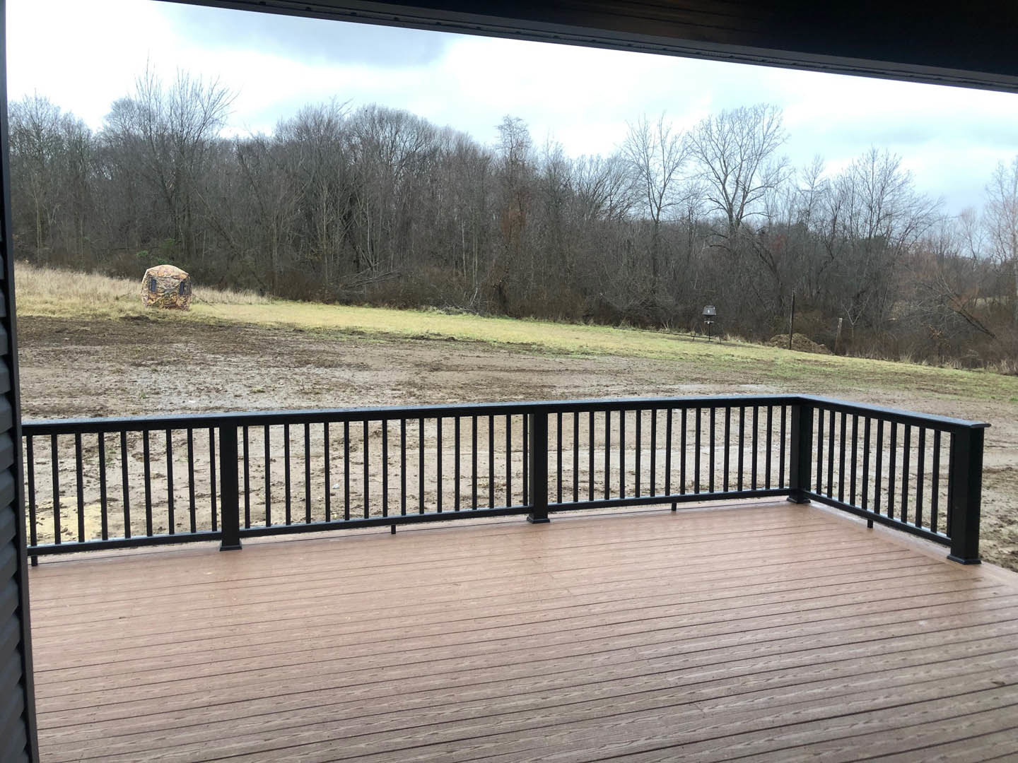 Wood deck with black metal railing overlooking expansive dirt field, bordered by fence, with trees and sky in background