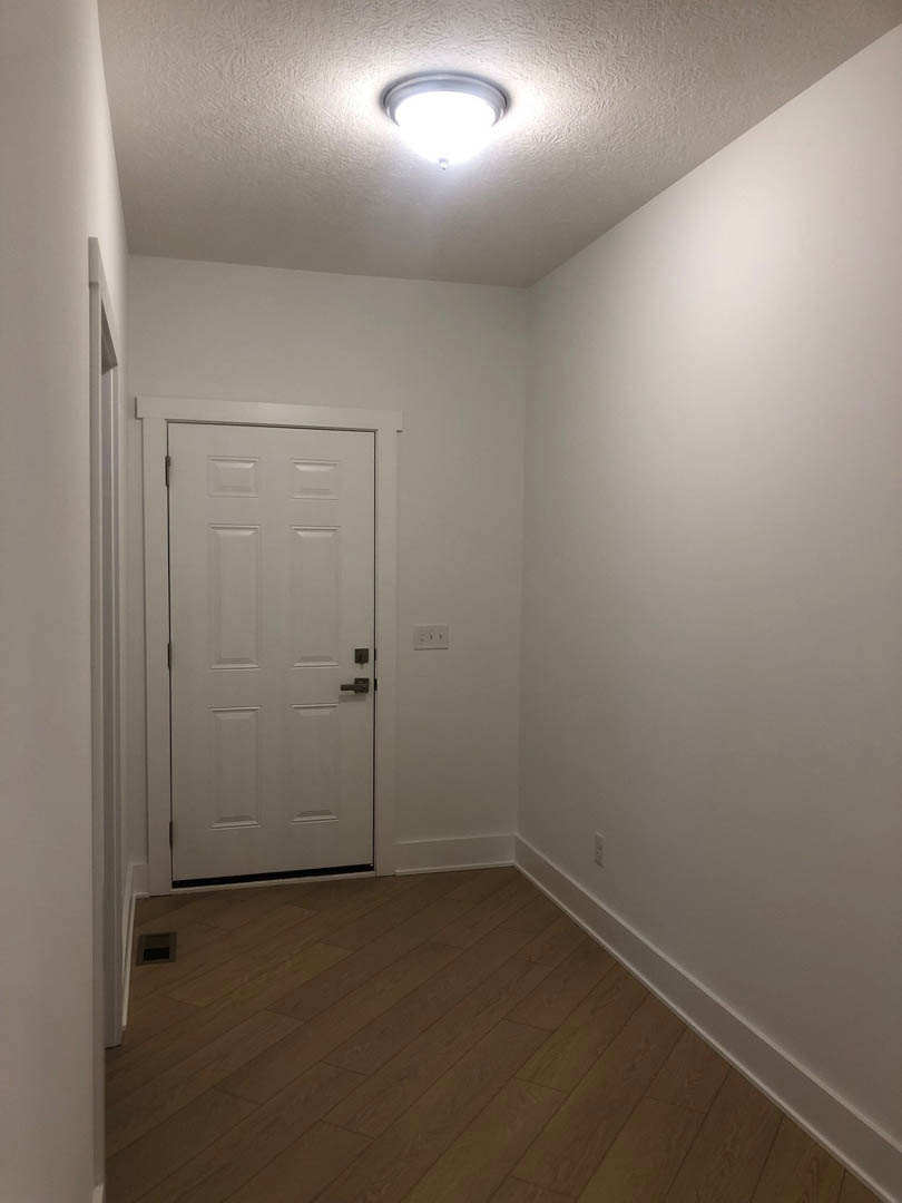 White door with black handle set in a room featuring wooden flooring, white baseboards, white walls with black trim, and a ceiling light fixture.