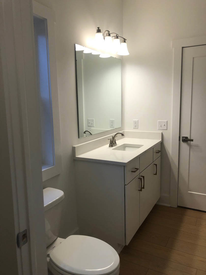 Modern bathroom featuring a rectangular mirror above a white sink with chrome faucet, grey countertop, closed white toilet, white walls, and a five-bulb light fixture.