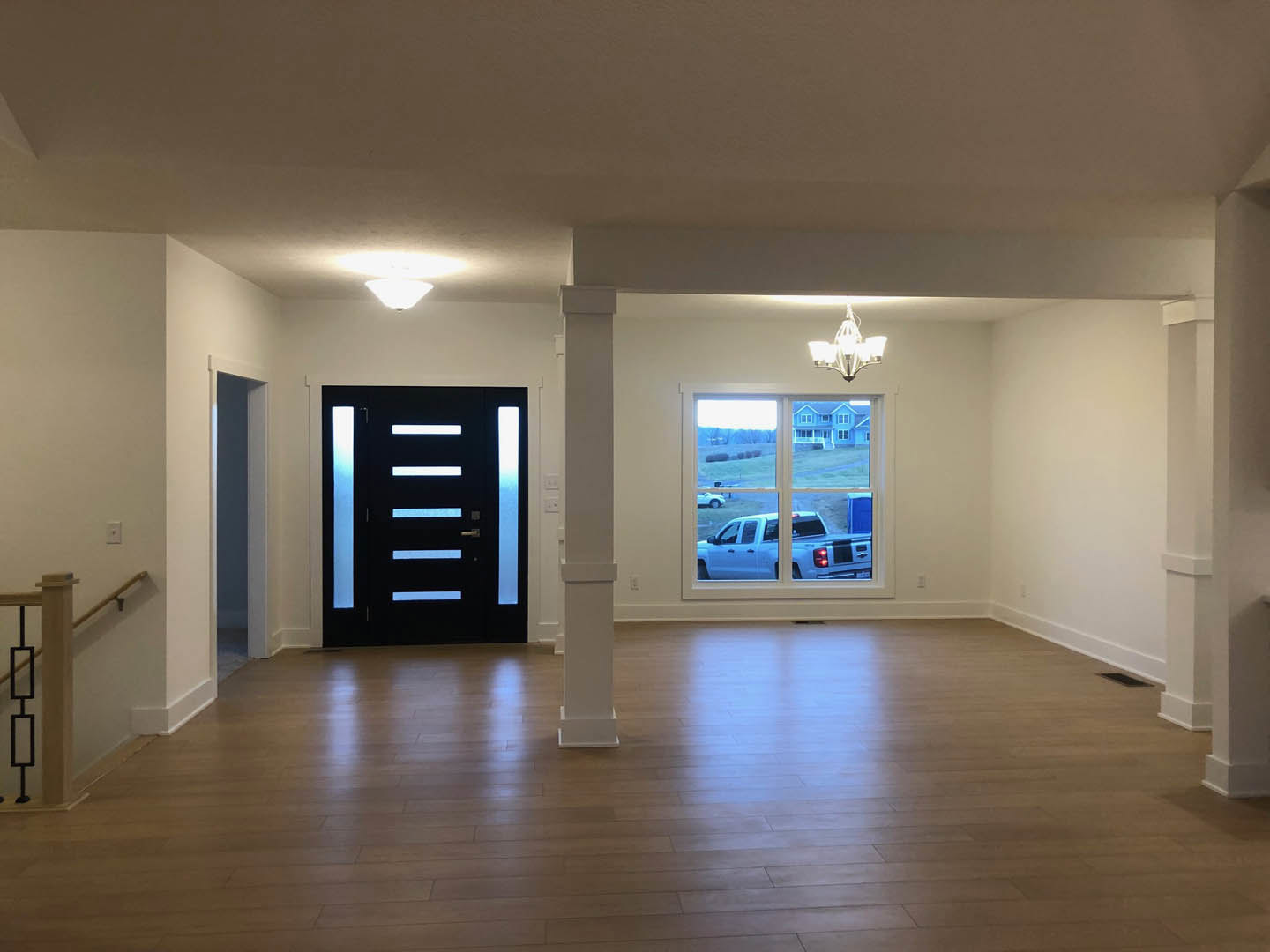 Black paneled door with frosted glass, wood laminate flooring, white column, five-light ceiling fixture, large window overlooking driveway with white truck parked outside