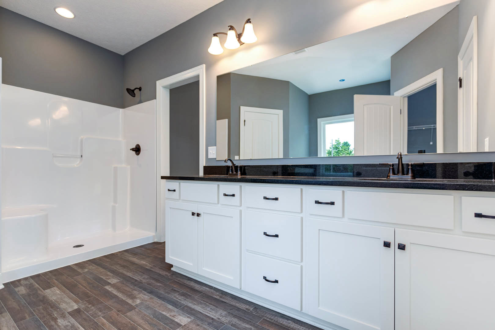 Bathroom with expansive mirror above white vanity, three-light fixture, white tiled shower with chrome showerhead, white door featuring black handle, white cabinetry with square