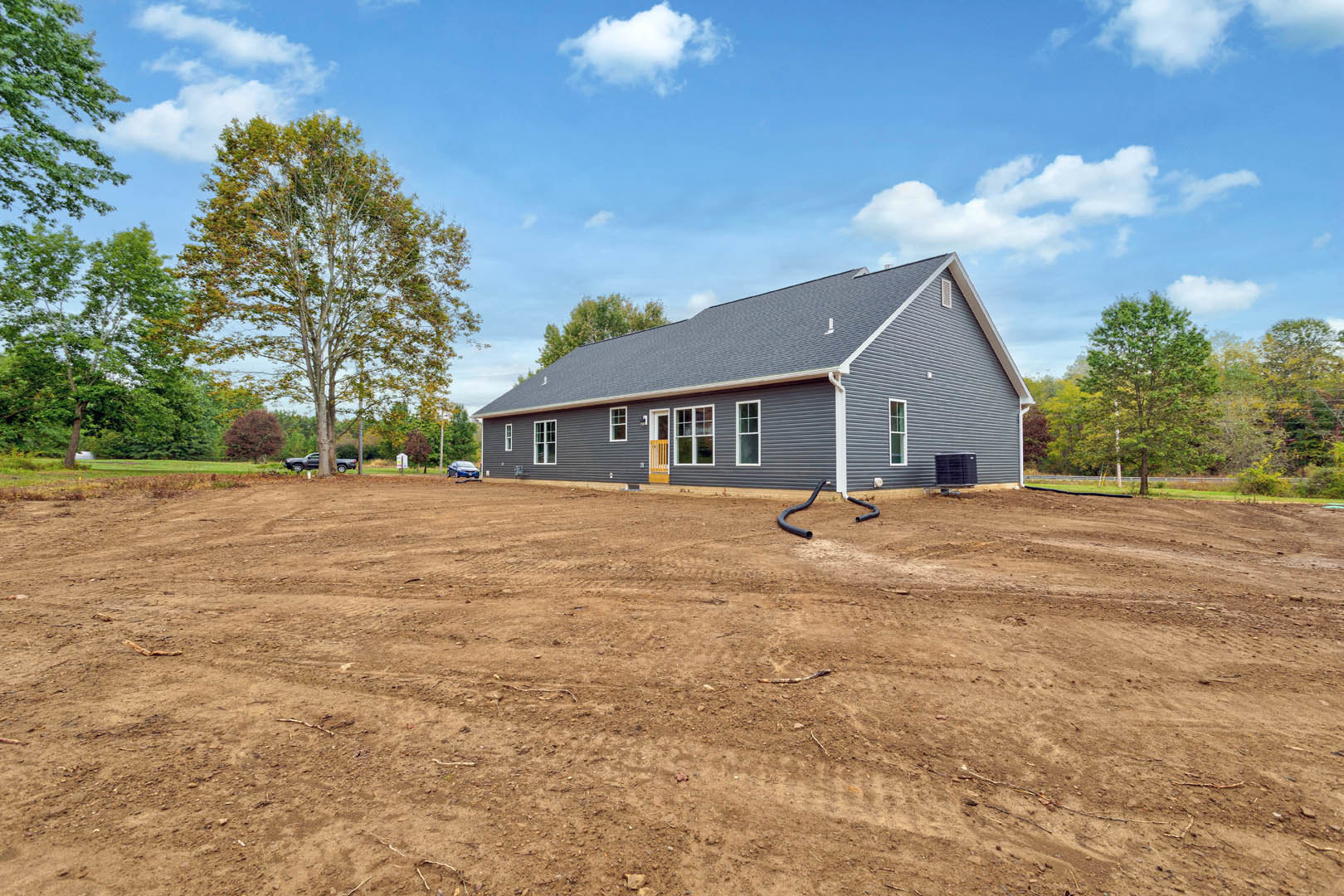 Modern cottage with yellow front door, surrounded by dirt lot, scattered black pipes, leafy trees, and a black square object on a pallet.