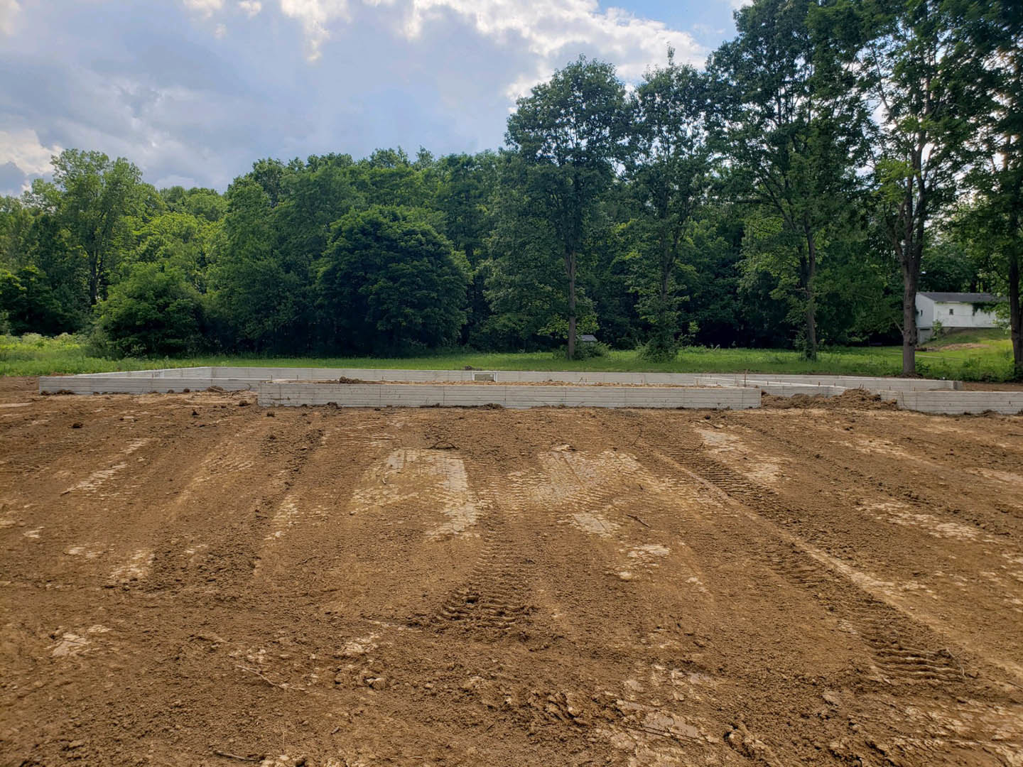 Dirt field with tire tracks bordered by a concrete wall, white building with window, and group of trees under blue sky