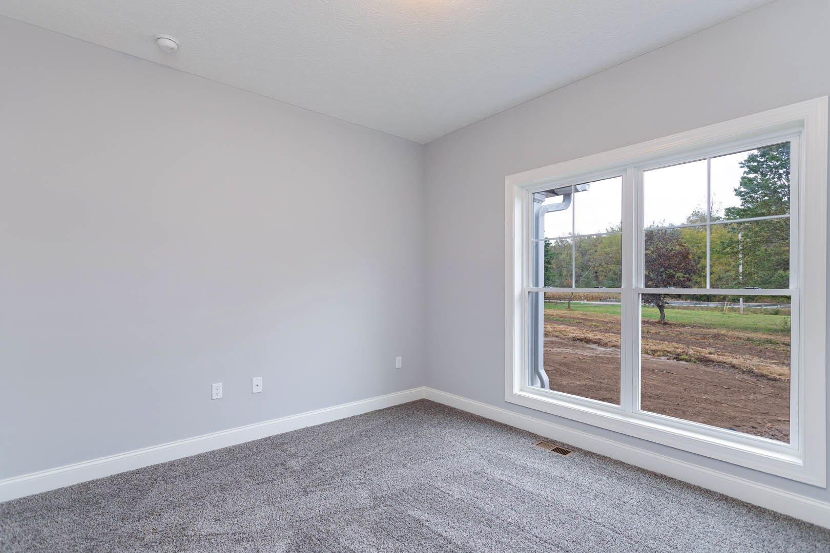 Carpeted room with white walls, large window overlooking dirt field, white ceiling with recessed light