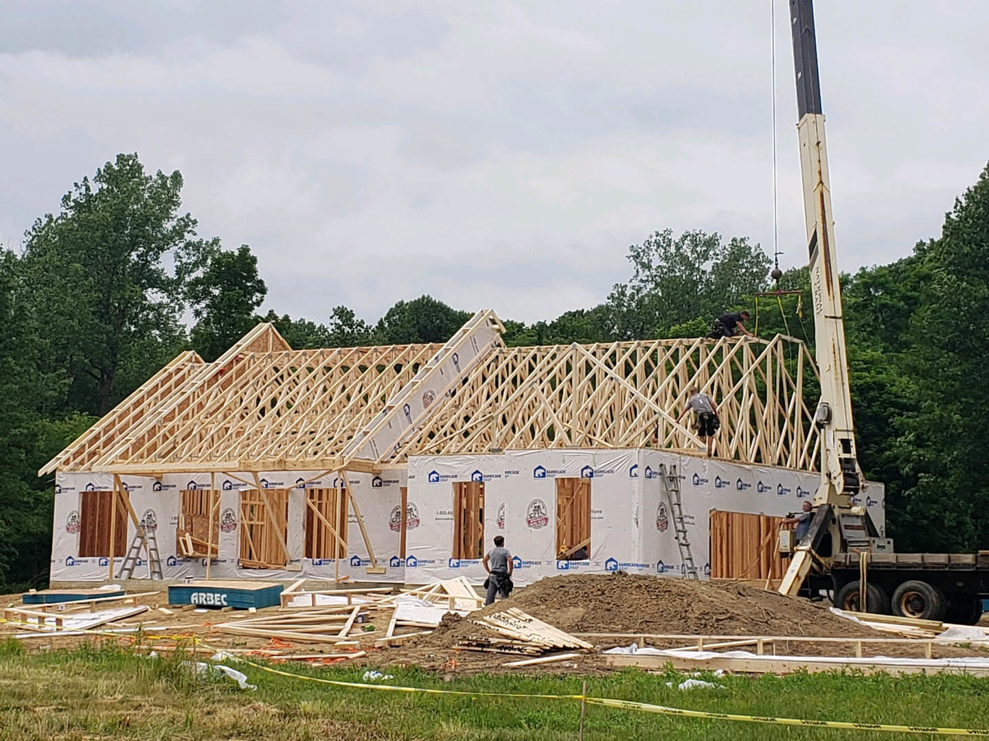 Framed house under construction with exposed wood beams, yellow caution tape on grass, people standing in front, trees and cloudy sky in background