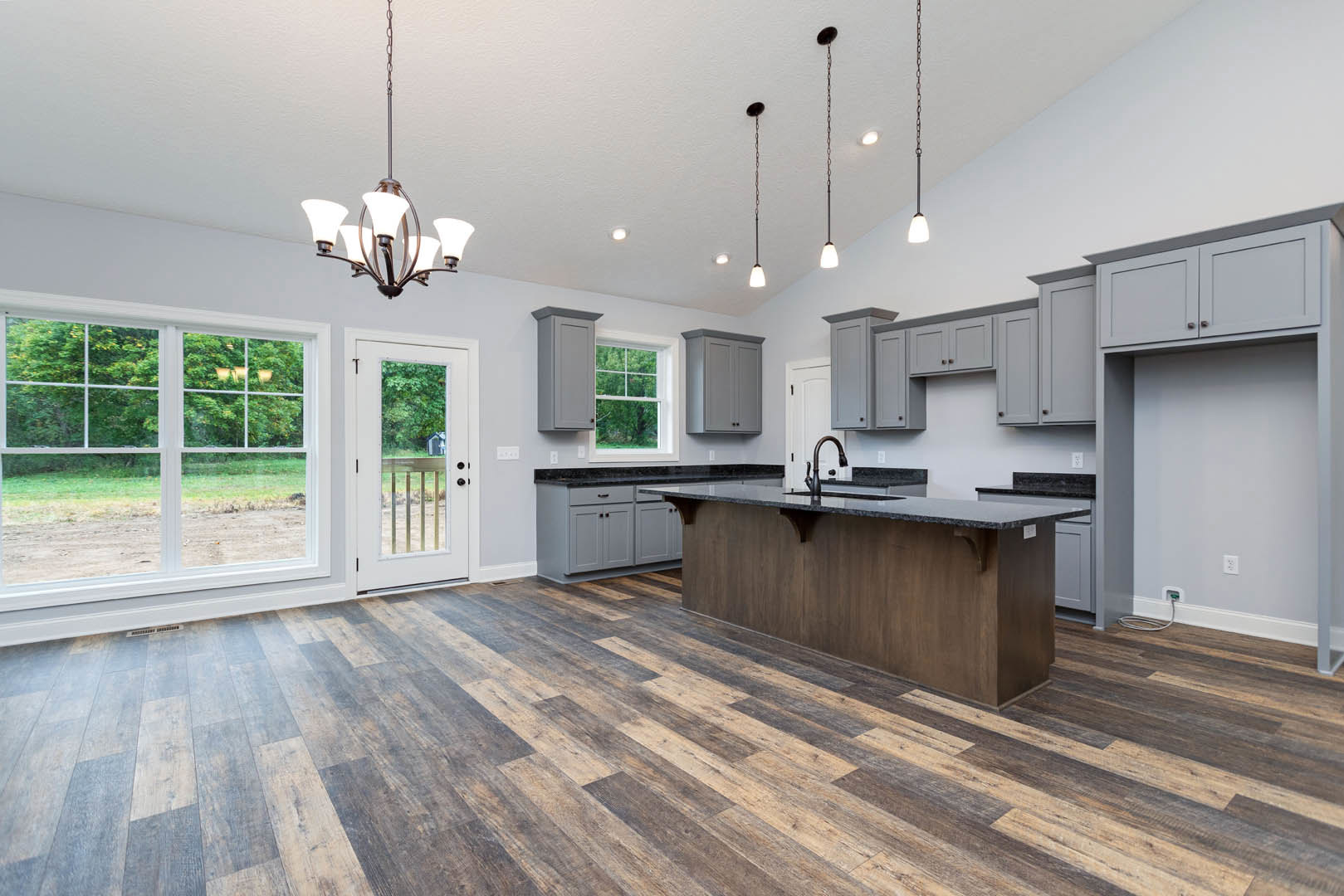 Spacious kitchen featuring a large island with black granite countertop, wood-grained flooring, white cabinetry, stainless steel sink, and a window overlooking a dirt road and