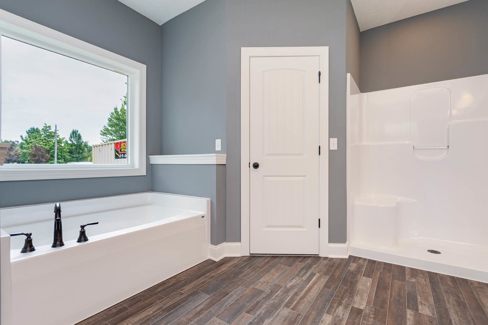 Bathroom with white shower stall, built-in bathtub with chrome faucets, wood flooring, white door with black knob, and window with white trim.