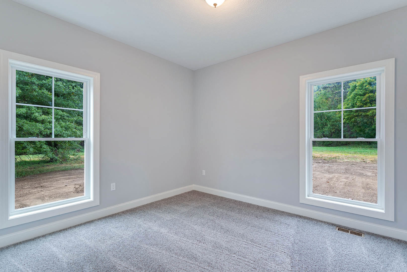 Carpeted room with large windows, white ceiling light, and views of trees and a dirt road outside