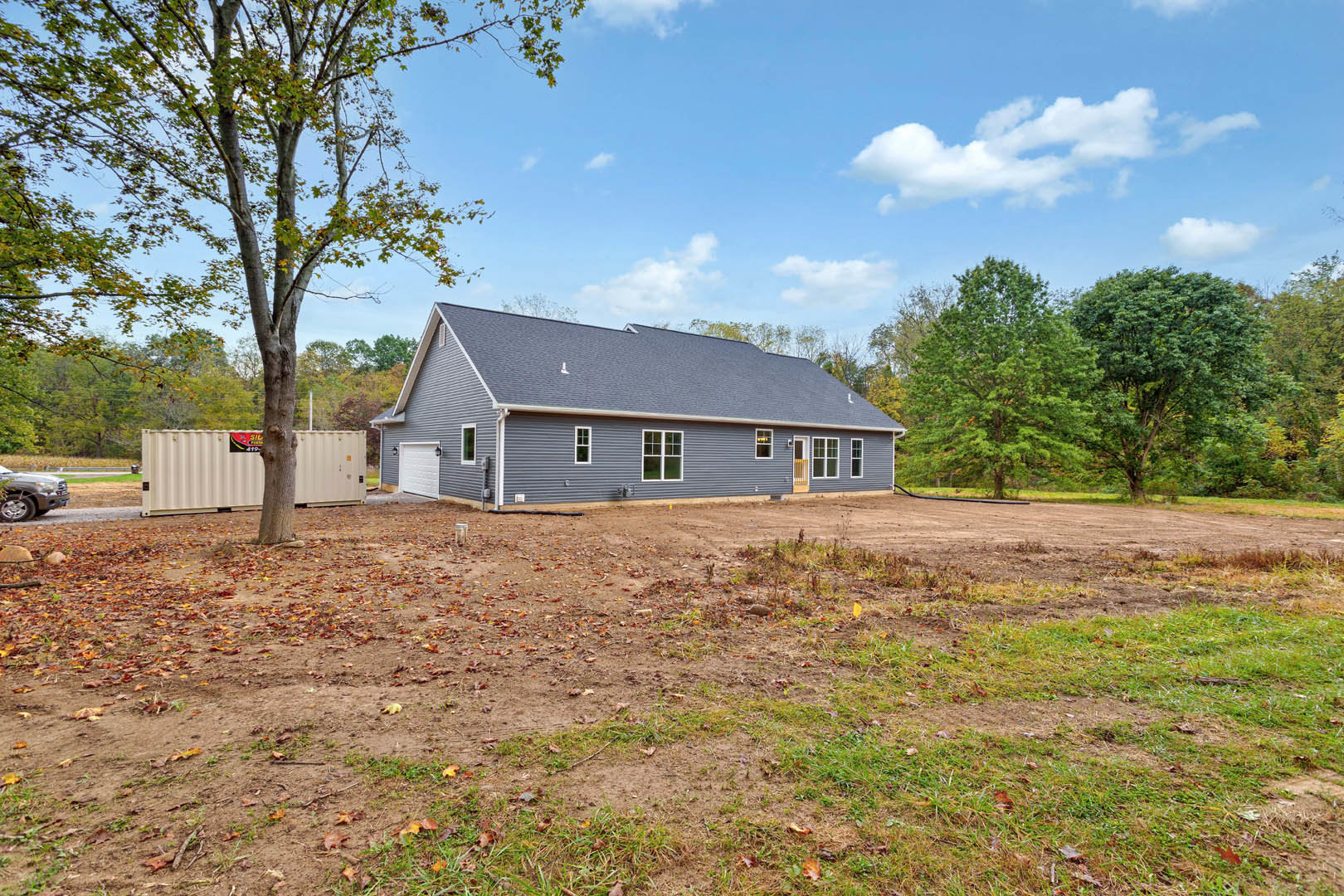 White cottage-style house with gray roof, surrounded by green grass yard and mature trees; metal container and tree trunk visible near side of house, scattered leaves on dirt patch