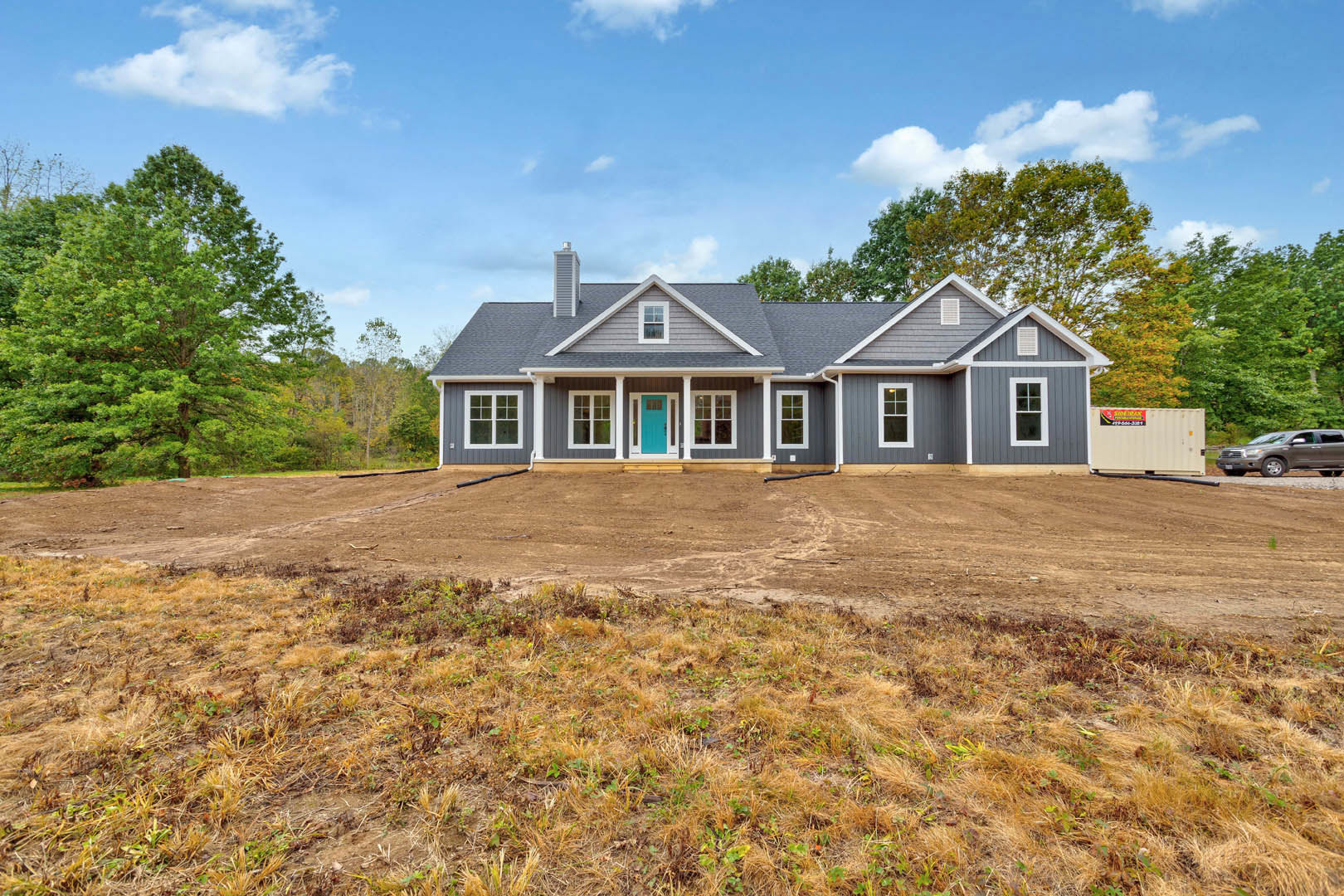 Single-story house with blue front door and white trim, surrounded by a dirt yard with patches of grass, white container with sign near driveway, car parked on adjacent road